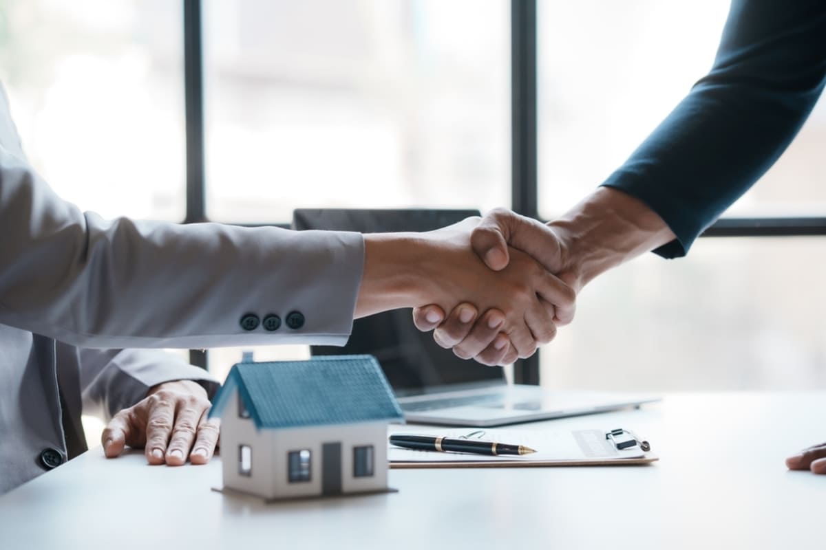 Two people shaking hands with a small home and a contract on a desk