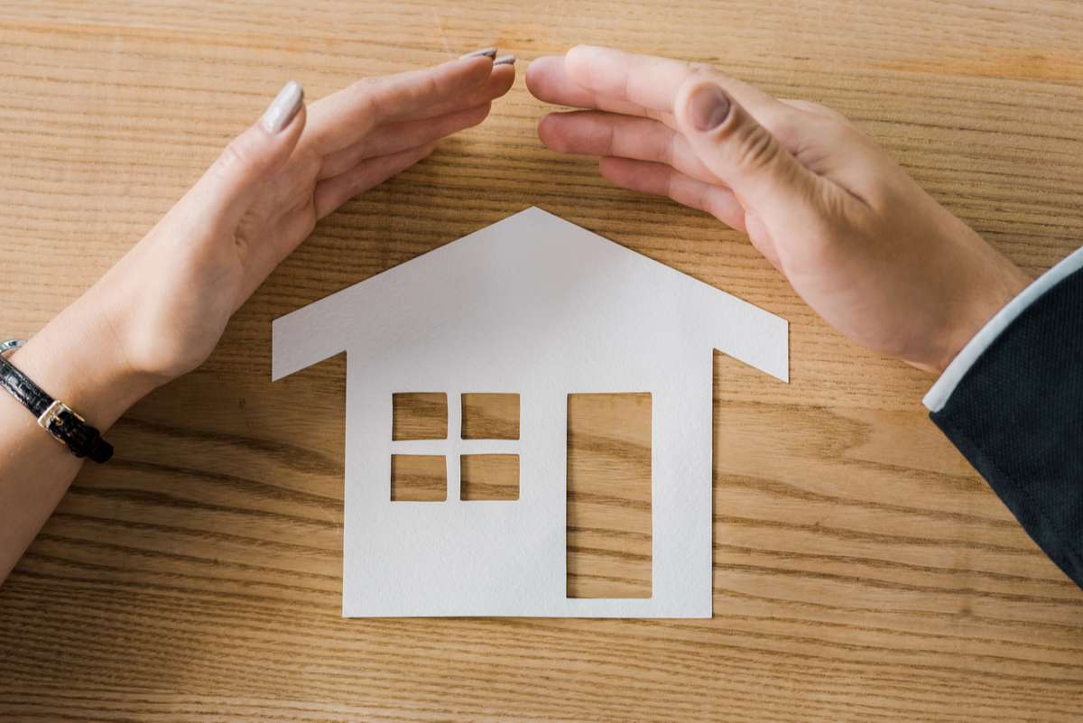 Partial view of business people covering house paper model on wooden tabletop with hands, insurance concept (R) (S)