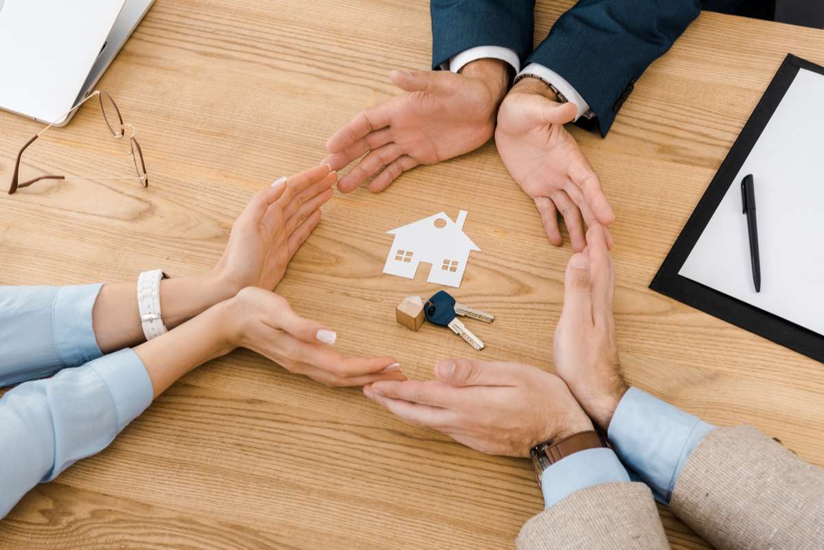 People making circle with hands on wooden table with paper house and keys inside, house insurance